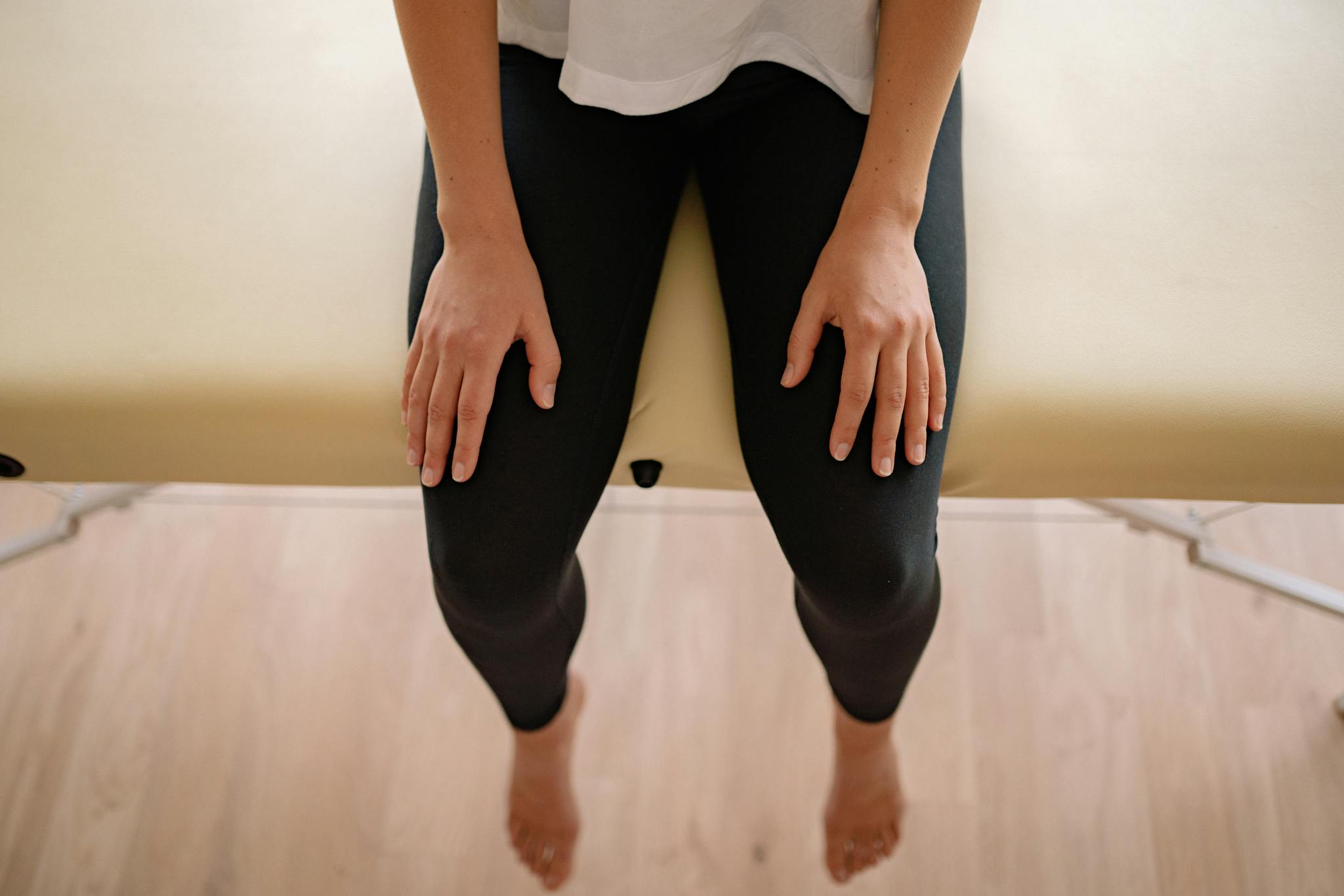 A woman sitting barefoot on a massage table indoors, captured from above.